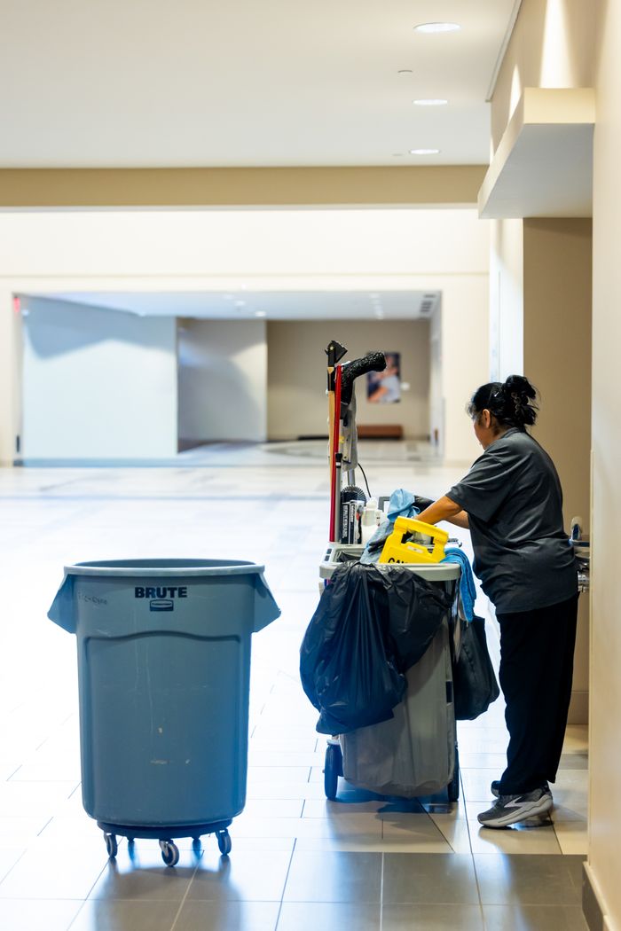 A janitorial worker pushes a cleaning cart in a spacious indoor hallway.